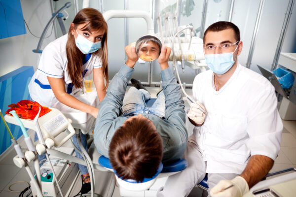 Young dentist doctor man in white uniform and special mask and young nurse woman examining man patient in dental office in clinic. Dental health and medical care concept