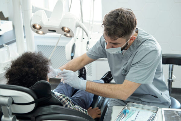 Dentist in uniform, glovea and protective mask bending over little boy while drilling his tooth after examination of oral cavity